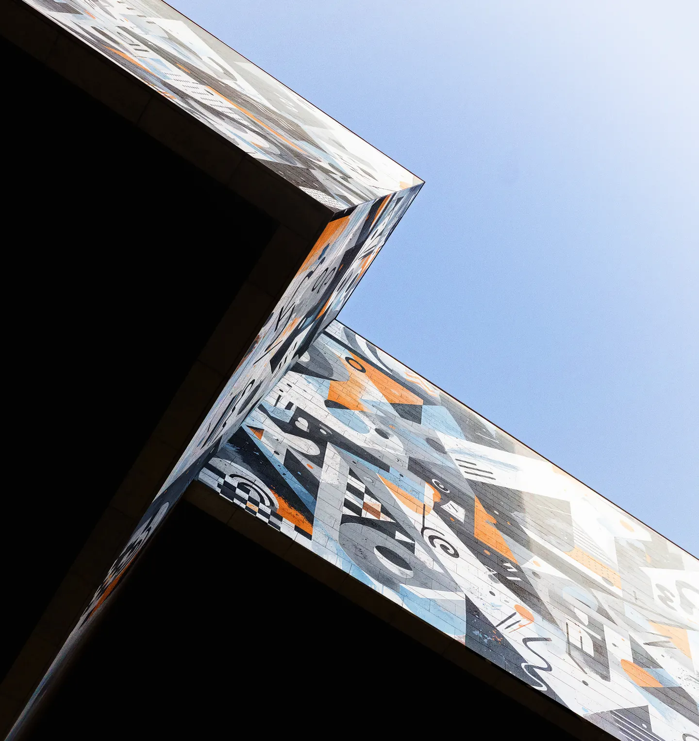 A building corner wrapped in a geometric mural of black, gray, light blue and orange shapes, photographed from below against a clear blue sky.